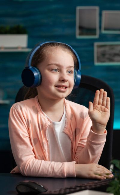 Little schoolchild wearing headset greeting remote teacher during online videocall meeting conference attenting to school lesson. Concept of distance education at home during coronavirus quarantine