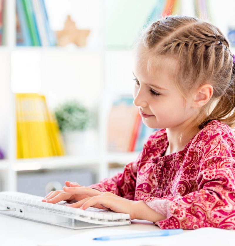 Little girl typing with fingers on computer keyboard sitting at desk in bright room